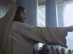 Man in a robe pointing forward in a dimly lit hall with pillars and seated people.